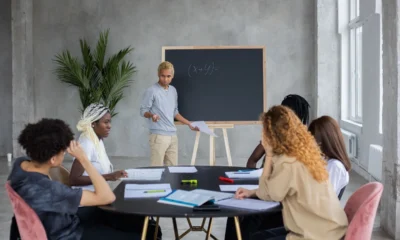Profesor explicando come si calcola il minimo comune multiplo en una pizarra durante una clase de matemáticas.